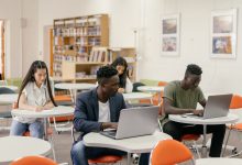 Estudantes usam laptops e fazem anotações durante uma atividade em uma sala de aula iluminada.