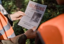 Mãos seguram um cartaz de pessoa desaparecida durante uma operação de busca em uma área de mata.