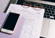 Laptop e smartphone ao lado de documentos fiscais sobre uma mesa de trabalho.