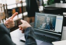Duas mulheres se comunicam por videochamada; uma delas sinaliza em Libras diante do laptop, em um ambiente de trabalho iluminado.