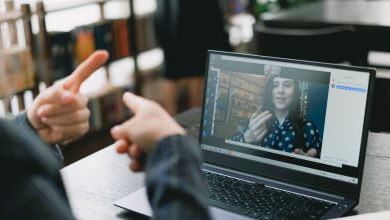 Duas mulheres se comunicam por videochamada; uma delas sinaliza em Libras diante do laptop, em um ambiente de trabalho iluminado.