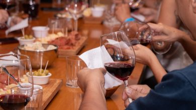Mãos seguram taças de vinho durante uma degustação, diante de petiscos dispostos sobre uma mesa de madeira.