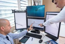Dois profissionais analisam dados financeiros exibidos em várias telas de computador em um escritório.
