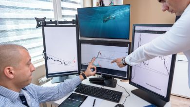 Dois profissionais analisam dados financeiros exibidos em várias telas de computador em um escritório.