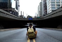 Homem sentado com uma mochila em uma via vazia sob um viaduto urbano.