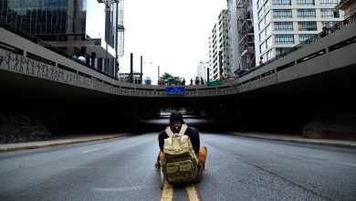 Homem sentado com uma mochila em uma via vazia sob um viaduto urbano.