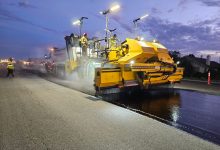 Trabalhadores operam máquinas pesadas durante a pavimentação de uma via ao entardecer.